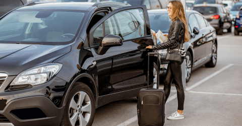 Jonge, vrolijke vrouw met een koffer en papieren in haar hand op een terrein waar auto's verhuurd worden